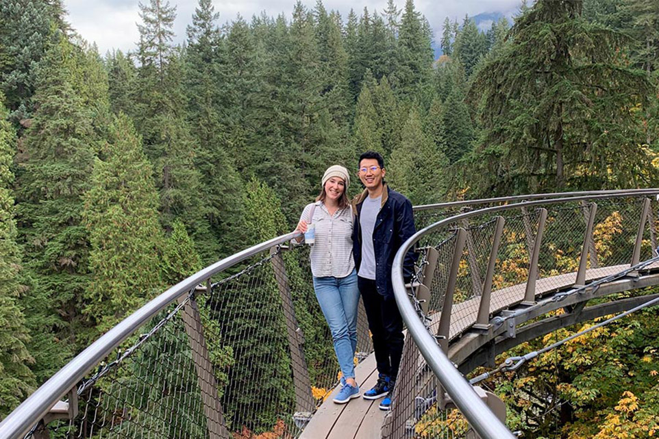 Woman and man stand on a suspended footbridge crossing a wooded area.