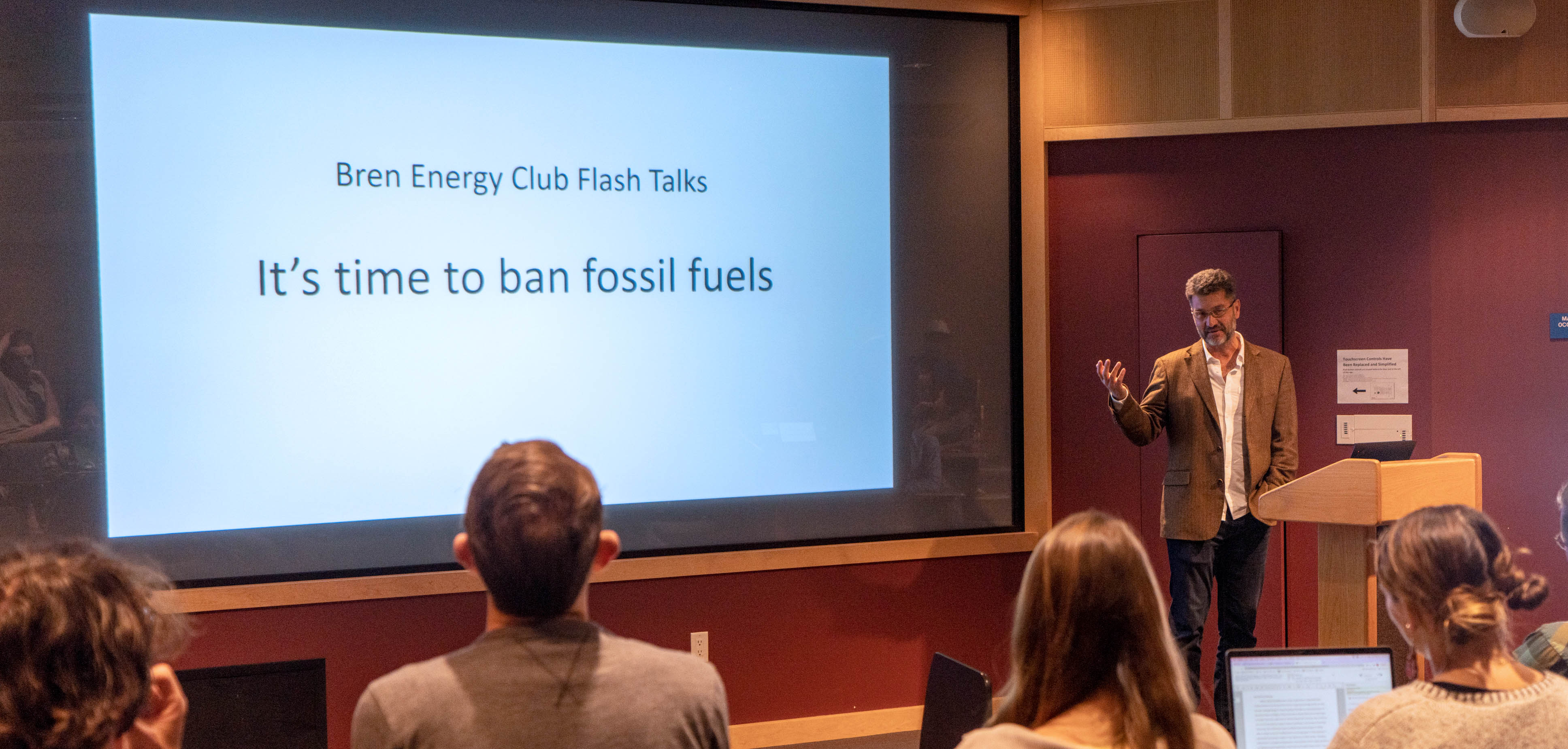 Professor Geyer giving a talk in lecture hall with projected slide "it's time to ban fossil fuels"