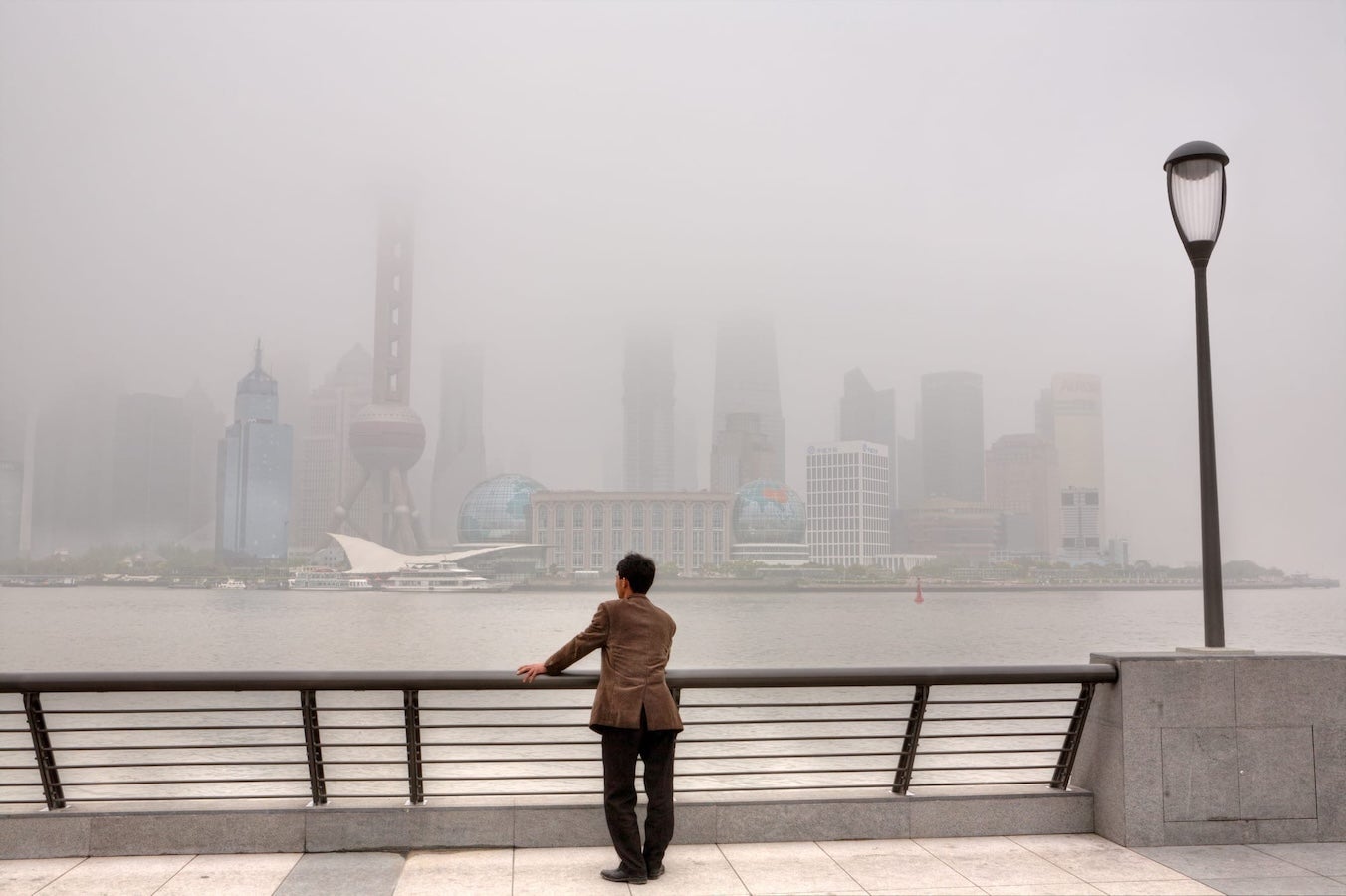 A person stands at a riverside railing looking toward a city skyline shrouded in heavy smog.