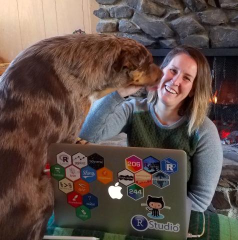 Woman on laptop and friendly dog by a fireplace