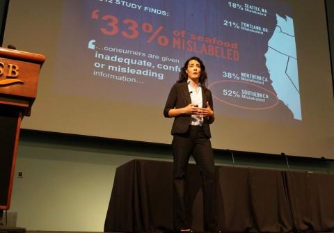 Woman presents on stage with projection on screen behind her