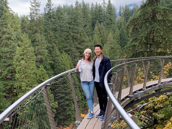 Woman and man stand on a suspended footbridge crossing a wooded area.