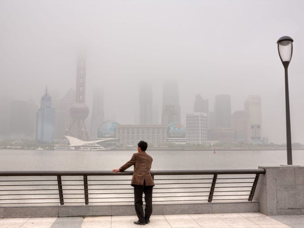  A person stands at a riverside railing looking toward a city skyline shrouded in heavy smog.