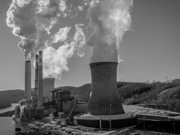 A large industrial power plant with multiple tall smokestacks emits thick plumes of steam or smoke into the sky. The facility sits beside a river, with wooded hills in the background. The scene is in black and white.