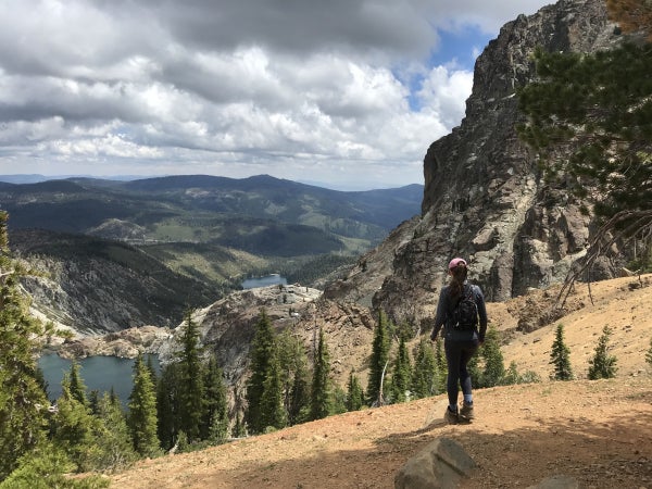 Alicia Magliato stands in front of a green landscape of trees, rocks, and a clouded sky.